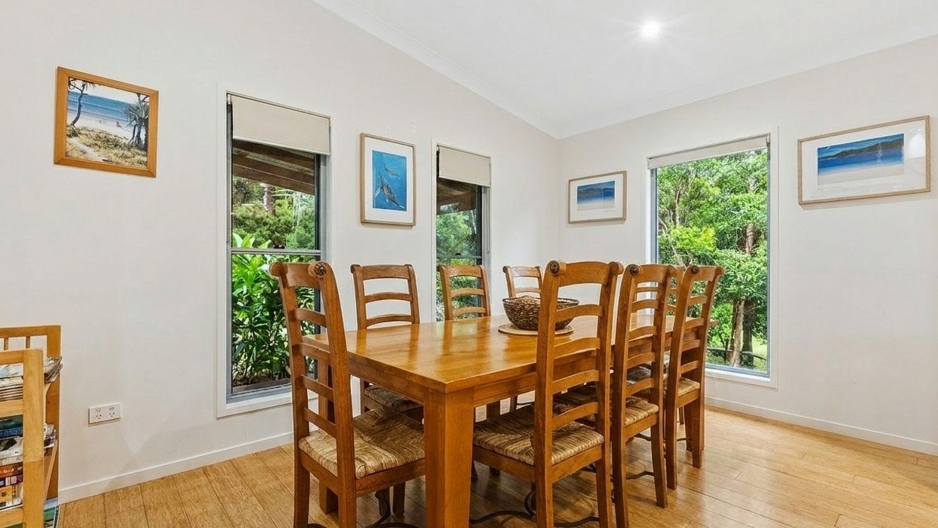 Un comedor con mesa y sillas de madera, ventanas amplias con vistas al follaje y cuadros en las paredes.