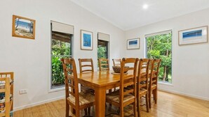 A dining room with a wooden table and chairs, large windows with views of greenery, and framed pictures on the walls. - 15 Esprit Drive - Rainbow Shores. Wifi. Close to beach (Rainbow Beach)