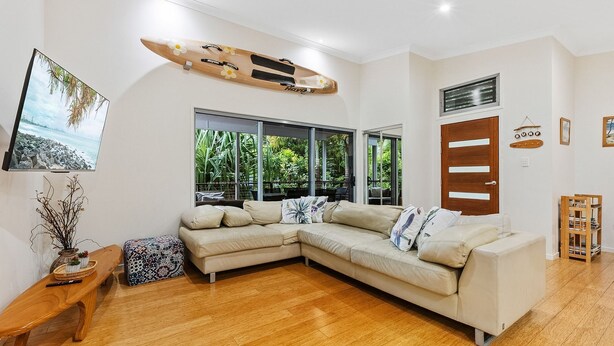 A living room with a sectional sofa, a wooden coffee table, a TV mounted on the wall, and a large window with a view of greenery. - 15 Esprit Drive - Rainbow Shores. Wifi. Close to beach (Rainbow Beach)