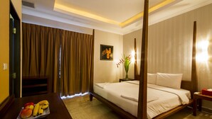 A hotel room with a four-poster bed, a wooden desk with fruit, a vase with flowers, and a framed picture on the wall.