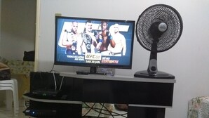 Un téléviseur diffusant un événement de l’UFC 220, un meuble noir pour appareils électroniques et un ventilateur sur pied.