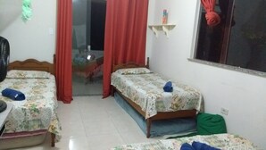 Une chambre avec deux lits, une fenêtre, un ventilateur et un sac vert posé sur le sol.