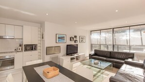 A modern living room with a glass coffee table, a leather sofa, a flat-screen TV, and a kitchen with white cabinetry and appliances.