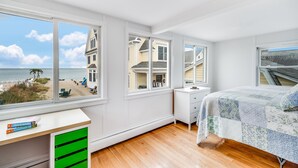 Een slaapkamer met een groot raam dat uitkijkt op een strand, een bed met quilt, een bureau met boeken en een dressoir.