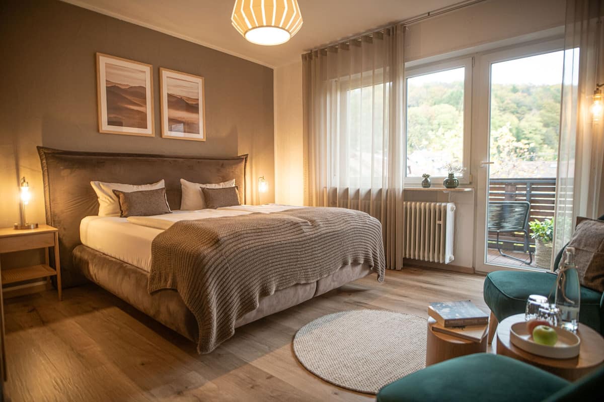Une chambre avec un grand lit, une table de chevet, un tapis rond, un fauteuil vert et une vue sur la verdure par la fenêtre.