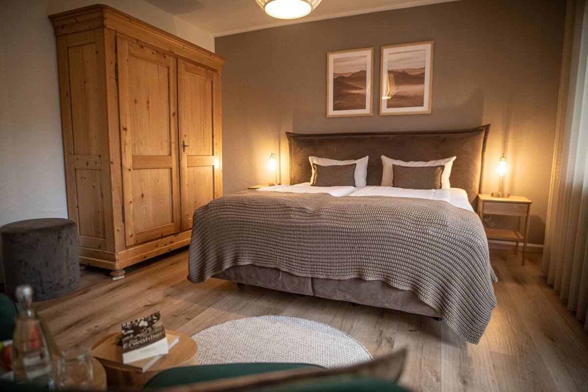 Une chambre avec une grande armoire en bois, un lit recouvert d’une couvre-lit à motifs, deux cadres photos accrochés au mur et un tapis rond posé sur le plancher en bois.