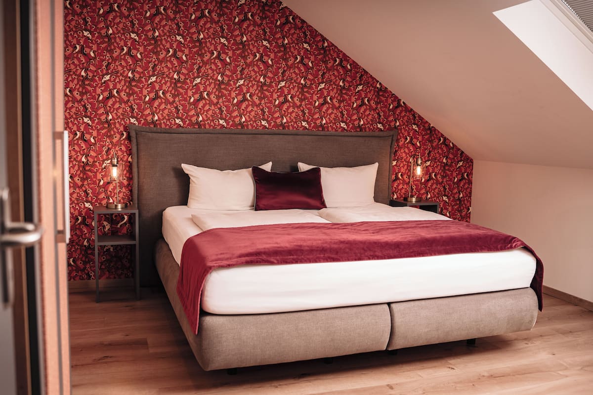 Une chambre avec un plafond en pente, un lit avec une tête de lit, un mur décoré d’un motif rouge et deux lampes de chevet.