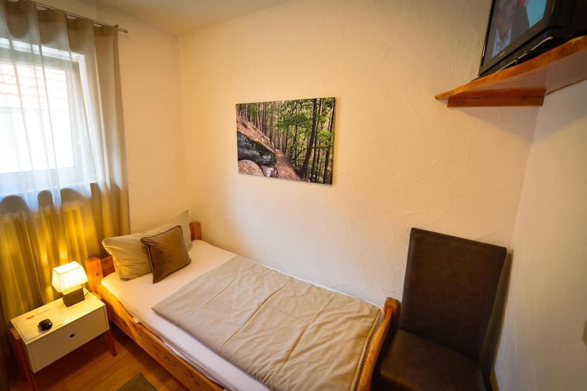 Une petite chambre à coucher, bien rangée, avec un lit simple, une table de chevet avec une lampe, une chaise et un tableau accroché au mur.
