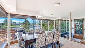 A spacious dining area with a large table and chairs, large windows offering a view of the outdoors, and a balcony with seating. - Shearwater House - Riverfront Accommodation (Port Fairy)