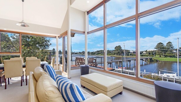 A spacious living area with large windows overlooking a body of water, a dining area with chairs and a table, and a modern interior design. - Shearwater House - Riverfront Accommodation (Port Fairy)