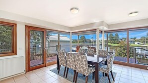 A dining area with a table and chairs, large windows with a view of a residential area and a body of water. - Shearwater House - Riverfront Accommodation (Port Fairy)