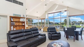 A spacious room with a large window offering a view of the outdoors, a wooden bookshelf, a leather sofa, and a dining area with chairs and a table. - Shearwater House - Riverfront Accommodation (Port Fairy)