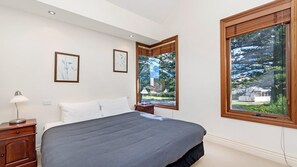 A bedroom with a large bed, a nightstand, a lamp, and two windows offering a view of the outdoors. - Shearwater House - Riverfront Accommodation (Port Fairy)