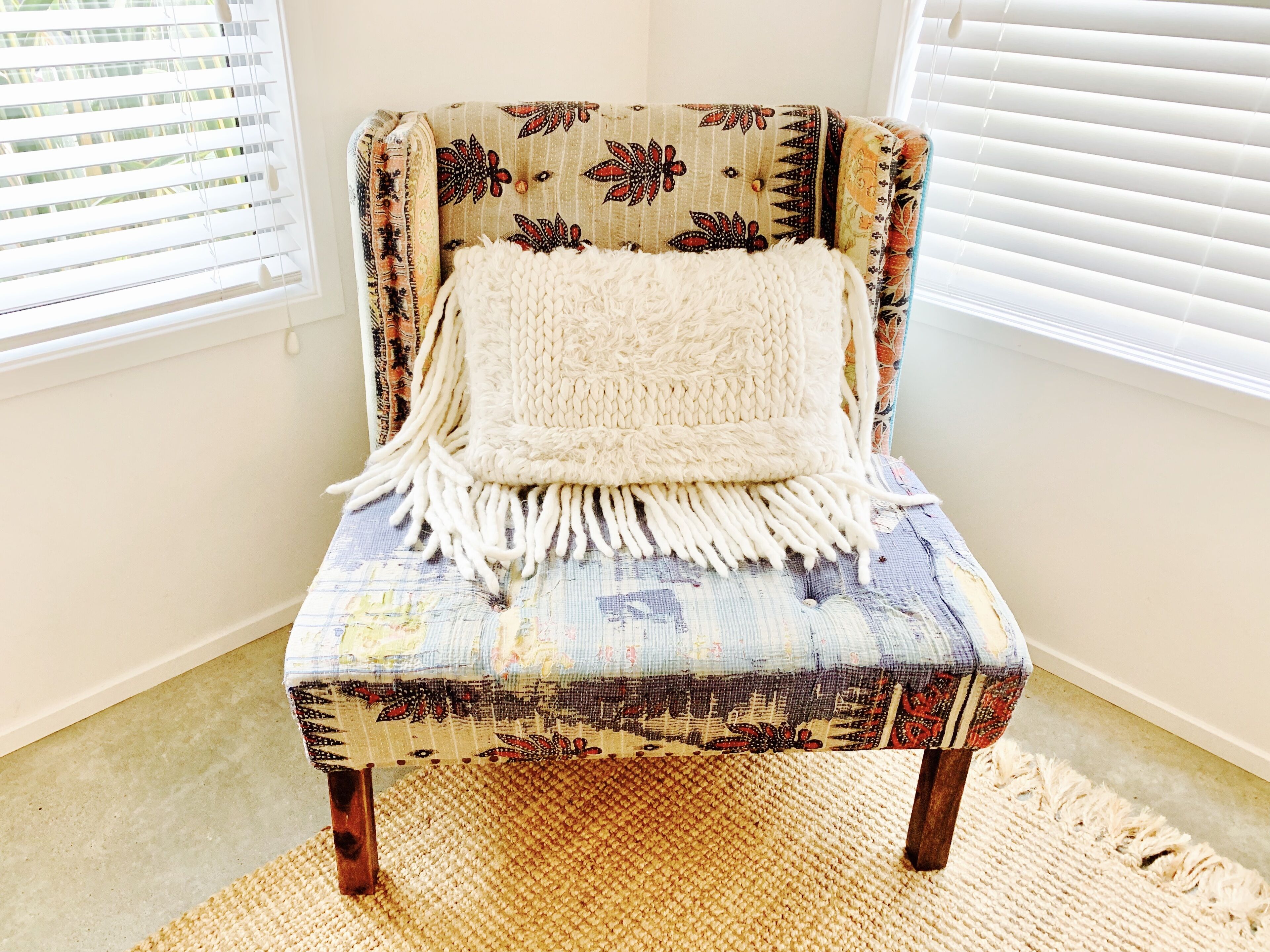 A chair with a patterned fabric and a white knitted throw, placed on a woven rug in a room with white walls and a window with blinds.
