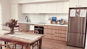 A modern kitchen with white cabinets, a stainless steel refrigerator, and a wooden dining table with chairs. - The Sea Life: 2 Min Walk to Beach + Private Outdoor Bath + Fireplace + Firepit (Manyana)