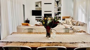A dining area with a wooden table, chairs, a vase with flowers, and a television in the background. - The Sea Life: 2 Min Walk to Beach + Private Outdoor Bath + Fireplace + Firepit (Manyana)