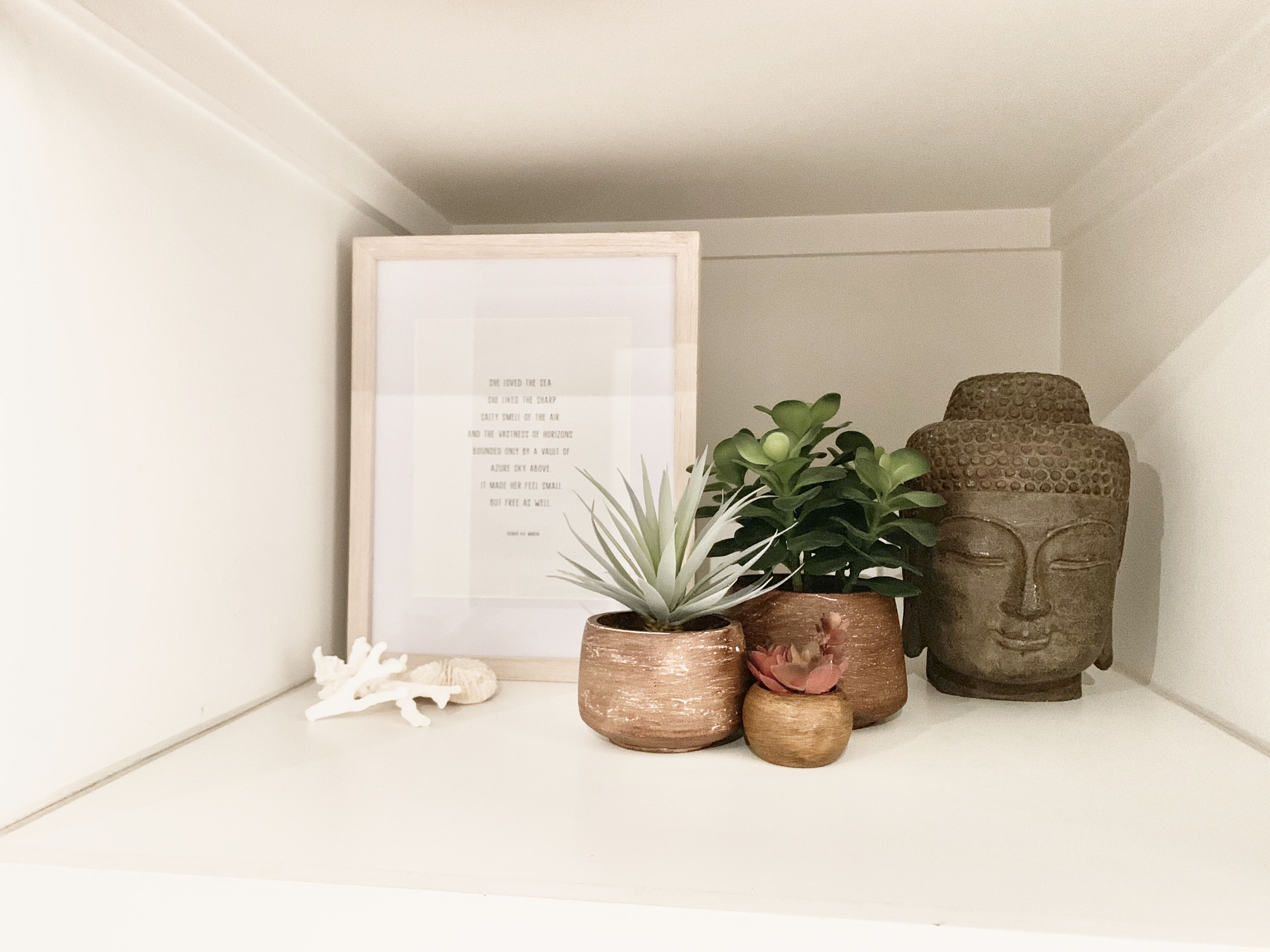 Un poema enmarcado sobre una estantería, dos plantas en macetas y una estatua de Buda.
