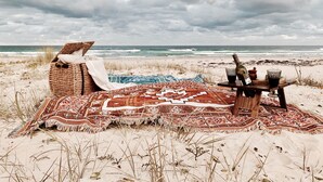 A beach with a wicker chair, a blanket, and a table with a bottle and glasses. - The Sea Life: 2 Min Walk to Beach + Private Outdoor Bath + Fireplace + Firepit (Manyana)