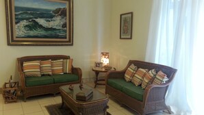 Un salon avec deux canapés en osier, une table basse, un tapis et un tableau représentant l’océan accroché au mur.