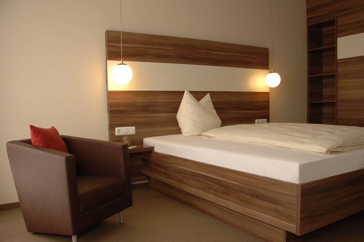 A modern hotel room with a wooden headboard, a single bed with white linens, a brown armchair with a red pillow, and two wall-mounted reading lamps.