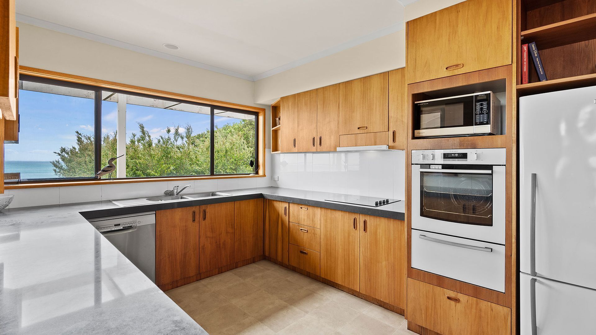 Une cuisine moderne avec des meubles en bois, un four encastré, un micro-ondes et une grande fenêtre donnant sur l’océan.