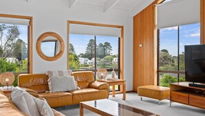 A modern living room with a leather sofa, a coffee table, and a TV stand. Large windows offer a view of the outdoors. - Beachfront - elevated ocean views (Port Fairy)