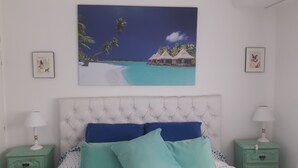 Una camera da letto con un'ampia tela raffigurante una spiaggia tropicale, una testiera trapuntata e due comodini con lampade.
