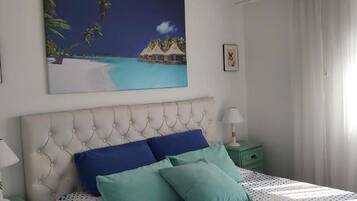 Un dormitorio con cabecera capitonada, cama con ropa de cama azul y verde azulado, dos mesitas de noche con lámparas, y un cuadro con una escena de playa en la pared.