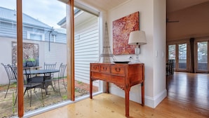 A room with a wooden floor, a dining table with chairs, a decorative cabinet, and a large window with a view of the outside. - Kinsale Cottage - perfect central location (Port Fairy)