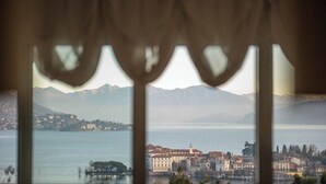 Vista a través de una ventana con cortinas, mostrando un lago, montañas y un pueblo con edificios.