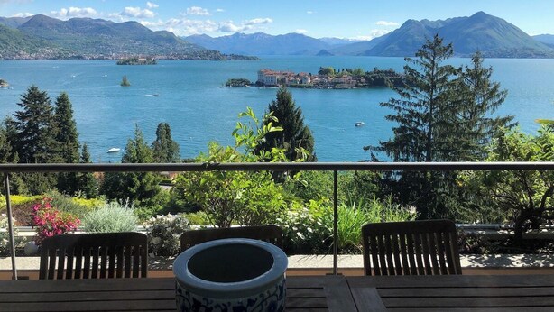 Un balcón con sillas y una mesa de madera, con vistas a un lago y una pequeña isla, con montañas al fondo.