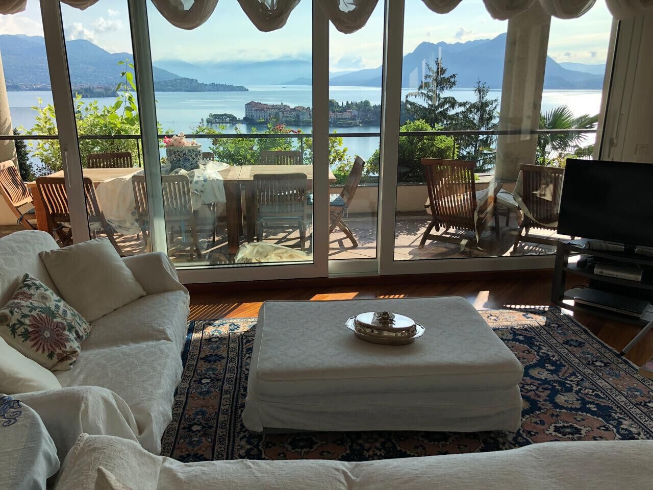 Ein Wohnzimmer mit Sofa, Hocker und Sesseln, einem Fernseher und Blick auf einen See und Berge durch große Fenster.