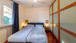 Una cama doble con cabecera, dos mesitas de noche con lámparas, un ventilador de techo, una ventana con cortinas y un radiador.
