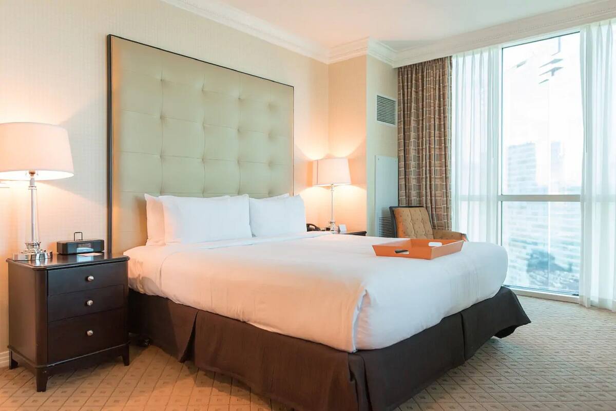 A hotel room with a large bed, two bedside tables with lamps, a view of the city through large windows, and a cityscape visible outside.