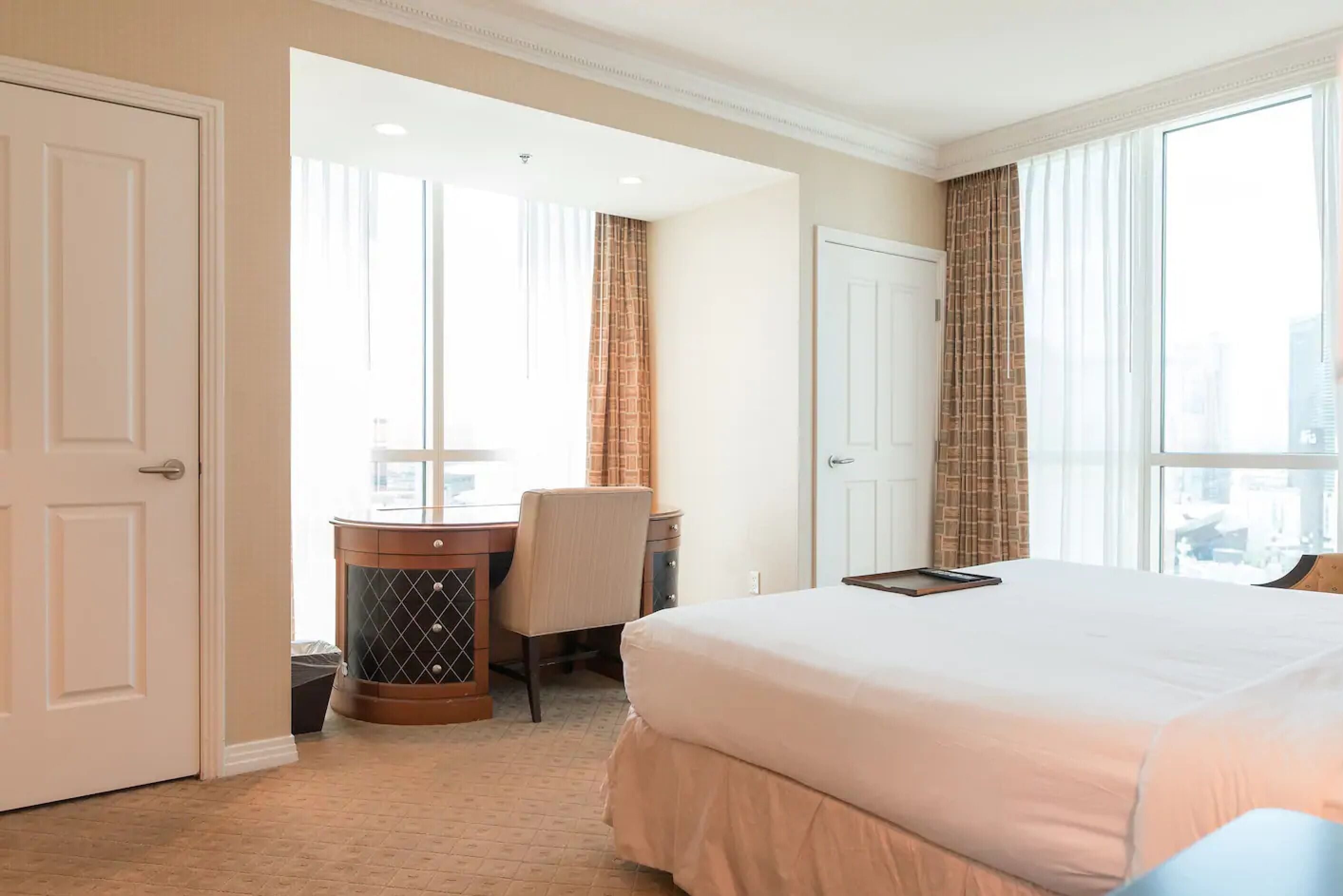Habitación de hotel con una cama grande, un escritorio y una silla. Se observan dos puertas y una ventana con cortinas.