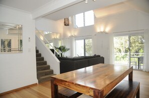 A spacious living area with a wooden table, a staircase, and a large window offering a view of the outdoors. - Byron Bay Beach Apartment BELONGIL BEACH (Byron Bay)