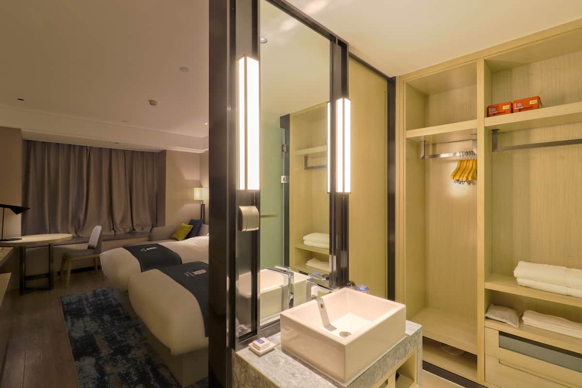A hotel room with a bed, bedside table, lamp, and a large mirror. There is a bathroom with a sink, mirror, and towel rack.