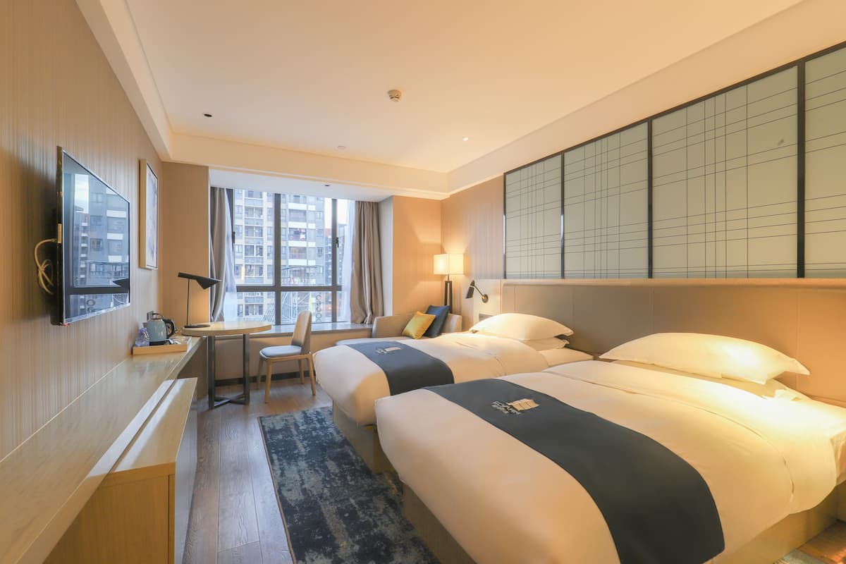A hotel room with two beds, a desk, a chair, a TV, and a large window with a city view.