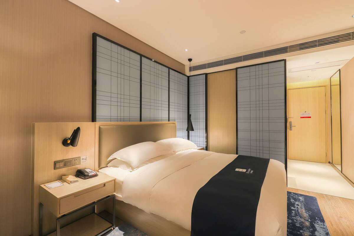 A modern hotel room with a large bed, a bedside table with a phone and a lamp, and a wardrobe with sliding doors.