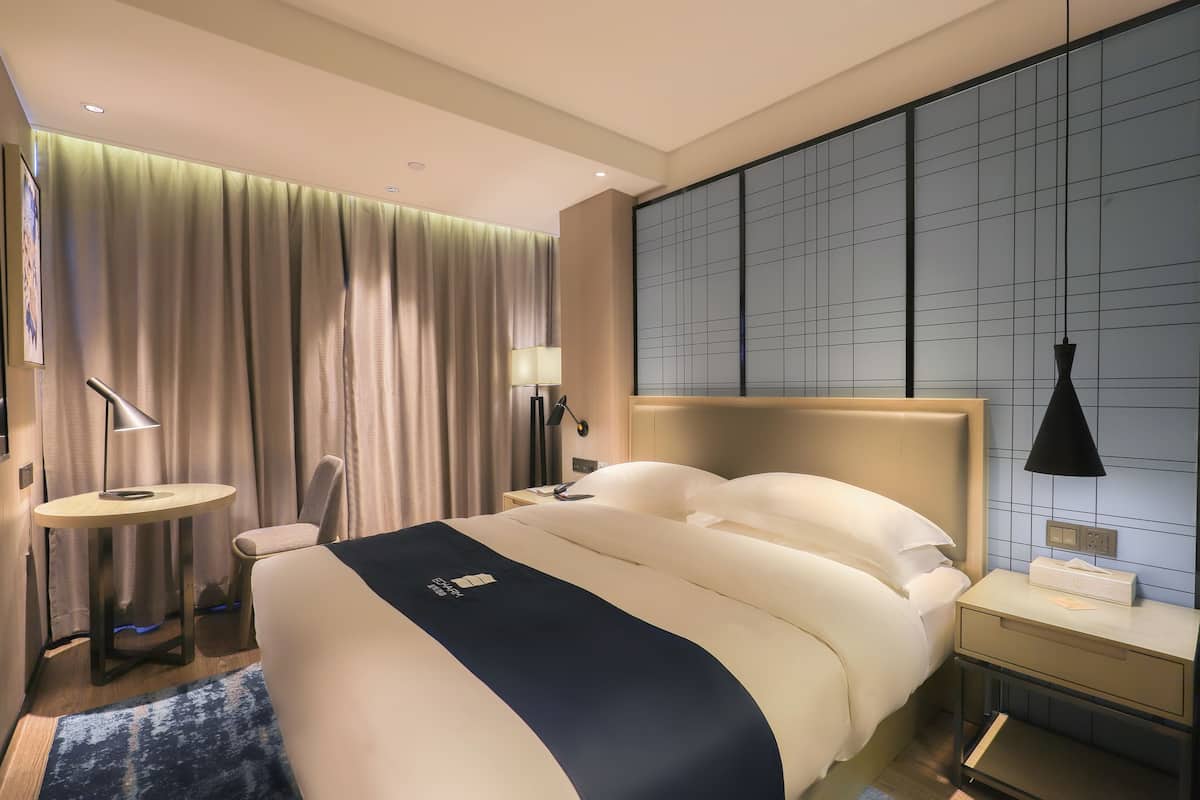 A hotel room with a large bed, a bedside table, a chair, a desk, and a large window with curtains.