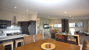 A modern kitchen with dark cabinets, a microwave, and a refrigerator. There is a dining area with a wooden table and wicker chairs. A television is mounted on the wall, and there is a fireplace with a stone surround. - Boronia - 7/30 Nettin Circuit (Jindabyne)