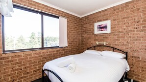 A bedroom with a brick wall, a bed with white linens, a window with curtains, and a framed picture on the wall. - Lodge 67 - 3/58 Townsend Street (Jindabyne)