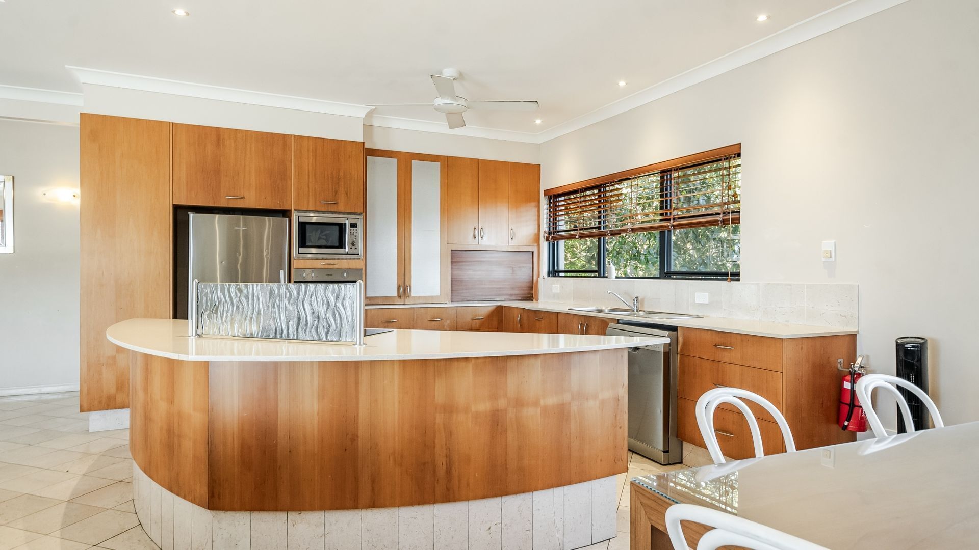 A modern kitchen with wooden cabinetry, a central island, stainless steel appliances, and a window with blinds.
