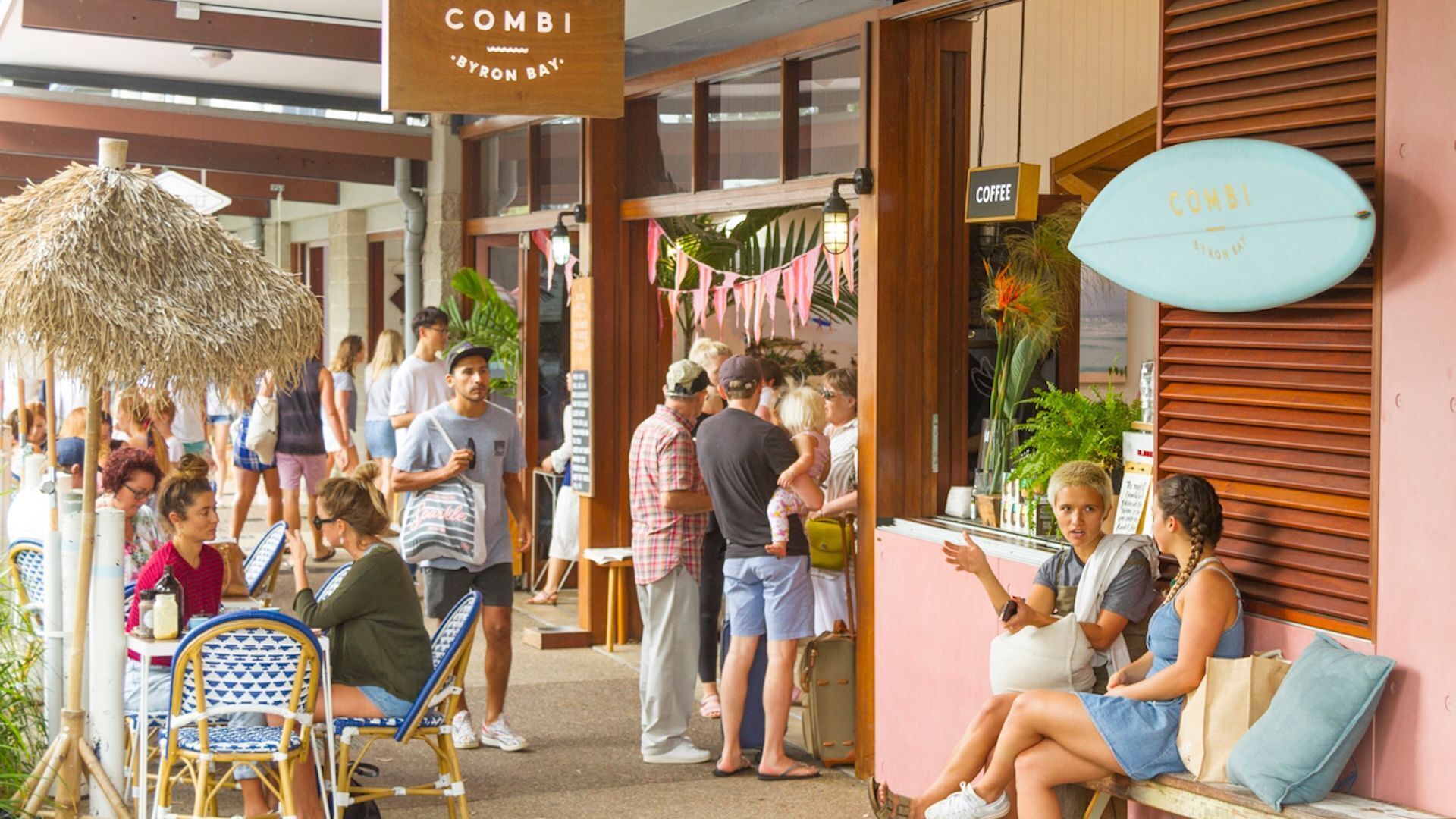 A bustling outdoor seating area with wicker chairs and tables, a coffee sign, and a surfboard with "COMBI" written on it.