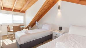 Een slaapkamer met een schuine kap, een bed, een nachtkastje met een lamp, een stoel, en een bureau met een stoel.