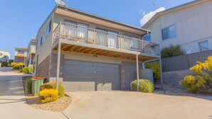 A two-story residential building with a covered garage, a balcony with metal railings, and a small garden area with shrubs and a tree.