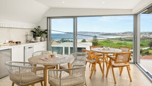 A modern kitchen with large windows, a dining area with wicker chairs, and a view of the sea and cityscape. - Luxury Beach Home Pool Ocean Views Walk to Beach (North Curl Curl)