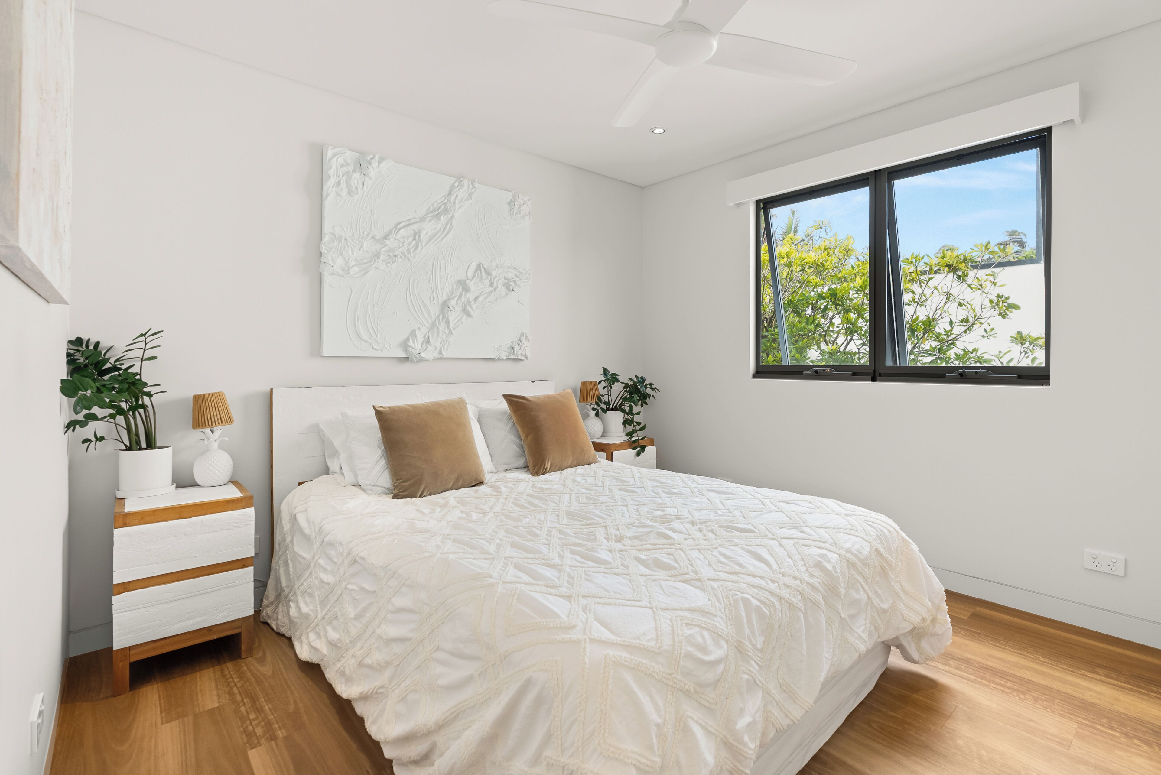 Une chambre dotée d’un grand lit, d’un ventilateur de plafond, d’une fenêtre donnant sur des arbres et d’une œuvre d’art accrochée au mur.