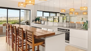 A modern kitchen with a large island, white cabinetry, and a view of the outdoors. - Luxury Beach Home Pool Ocean Views Walk to Beach (North Curl Curl)
