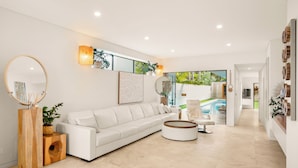 A modern living room with a white sofa, round coffee table, and wooden side tables. There is a large mirror, wall-mounted artwork, and a view of a pool through a glass door. - Luxury Beach Home Pool Ocean Views Walk to Beach (North Curl Curl)